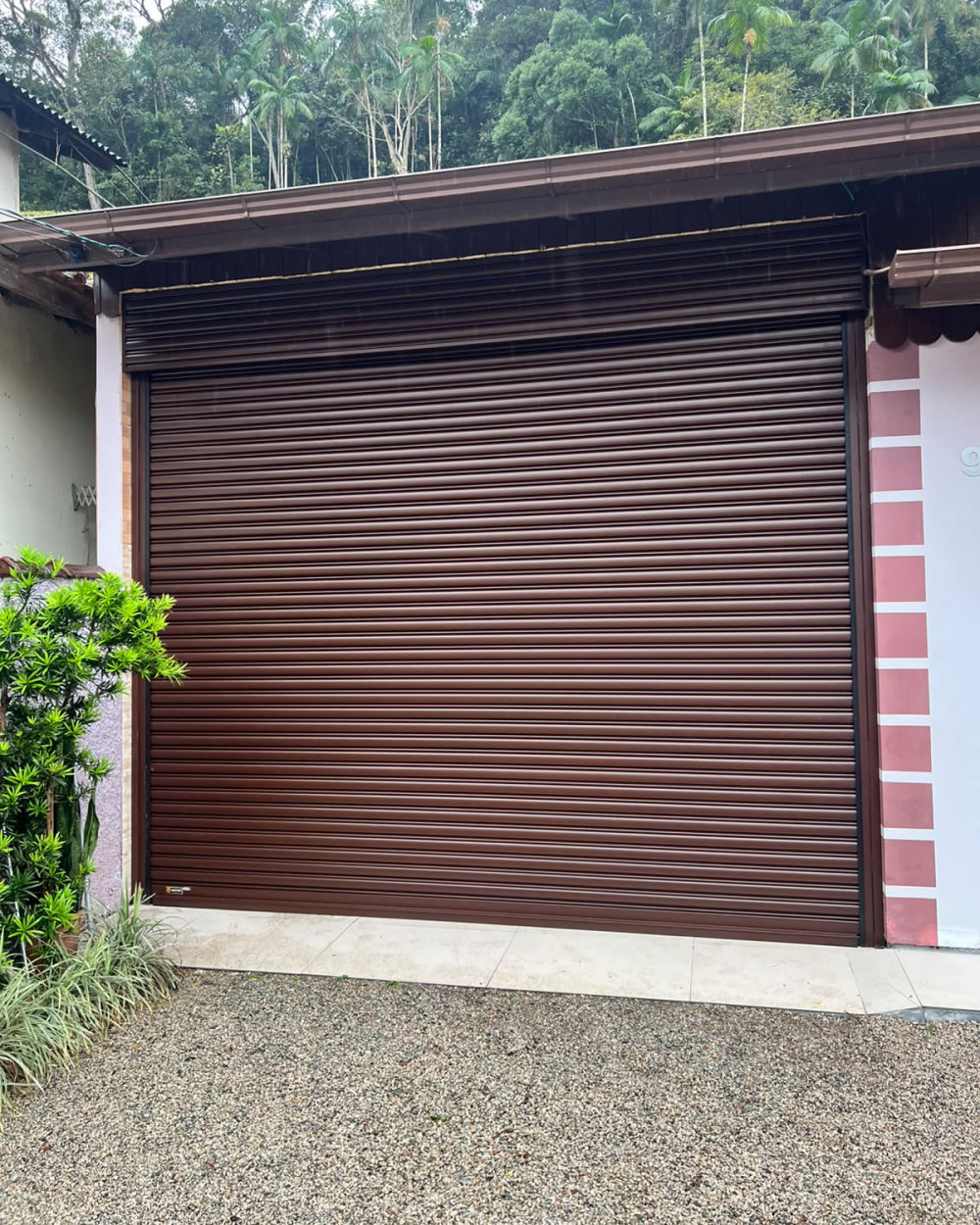 Portão de enrolar marrom instalado pela Pépe Serralheria na garagem de uma casa, com a mata ao fundo